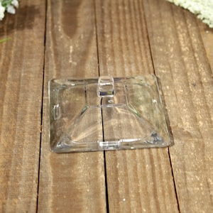 May include: A clear, square glass lid with a small handle. The lid has a slightly raised, pyramid-shaped base and is designed to fit a container. The image is taken from a top-down perspective.