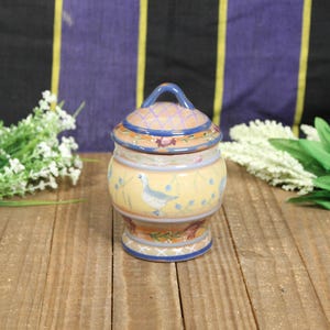 May include: A ceramic jar with a lid, featuring a yellow base with a duck design and blue accents. The lid has a blue handle and a patterned design. The jar is placed on a wooden surface with floral decorations.