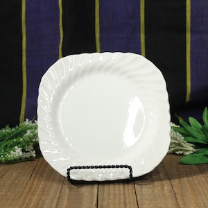 May include: A white, square-shaped plate with a swirled rim design, displayed on a black metal stand. The plate is set against a backdrop of a wooden surface and a patterned fabric.