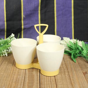May include: A vintage cream-colored condiment server with three compartments. The server has a yellow base and handle. The background includes a wooden surface, purple and black striped fabric, and artificial flowers.