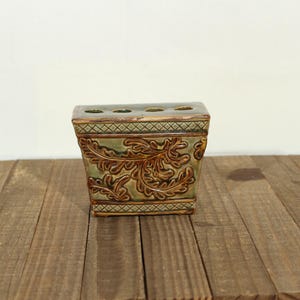 May include: A rectangular ceramic flower frog with a sage green and brown glaze. The exterior features a raised relief pattern of swirling acanthus leaves, bordered by a cross-hatched grid design at the top and bottom edges. It has four circular holes.