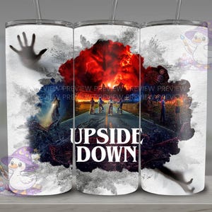 Pode incluir: Um copo branco com um design da série "Stranger Things". O design inclui uma explosão vermelha e preta, silhuetas de personagens e as palavras "UPSIDE DOWN" em branco. Uma mão e uma figura também são visíveis.
