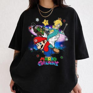 May include: Black t-shirt featuring a colorful graphic of Mario, Princess Peach, and other characters from the Super Mario Galaxy video game. The text "THE SUPER MARIO GALAXY" is displayed in a vibrant, multi-colored font.