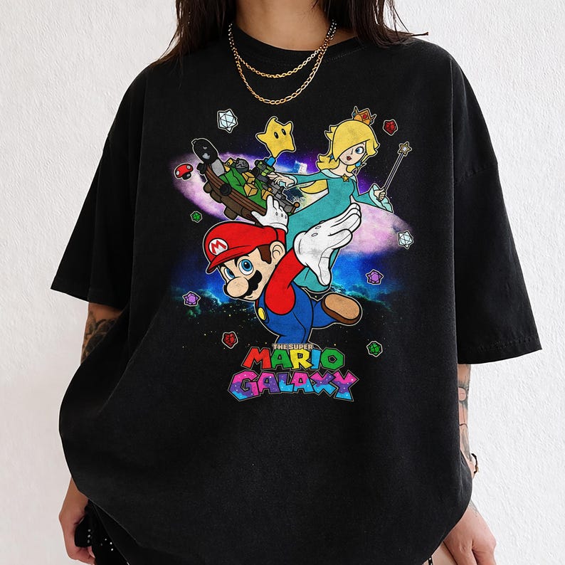 May include: Black t-shirt featuring a colorful graphic of Mario, Princess Peach, and other characters from the Super Mario Galaxy video game. The text "THE SUPER MARIO GALAXY" is displayed in a vibrant, multi-colored font.