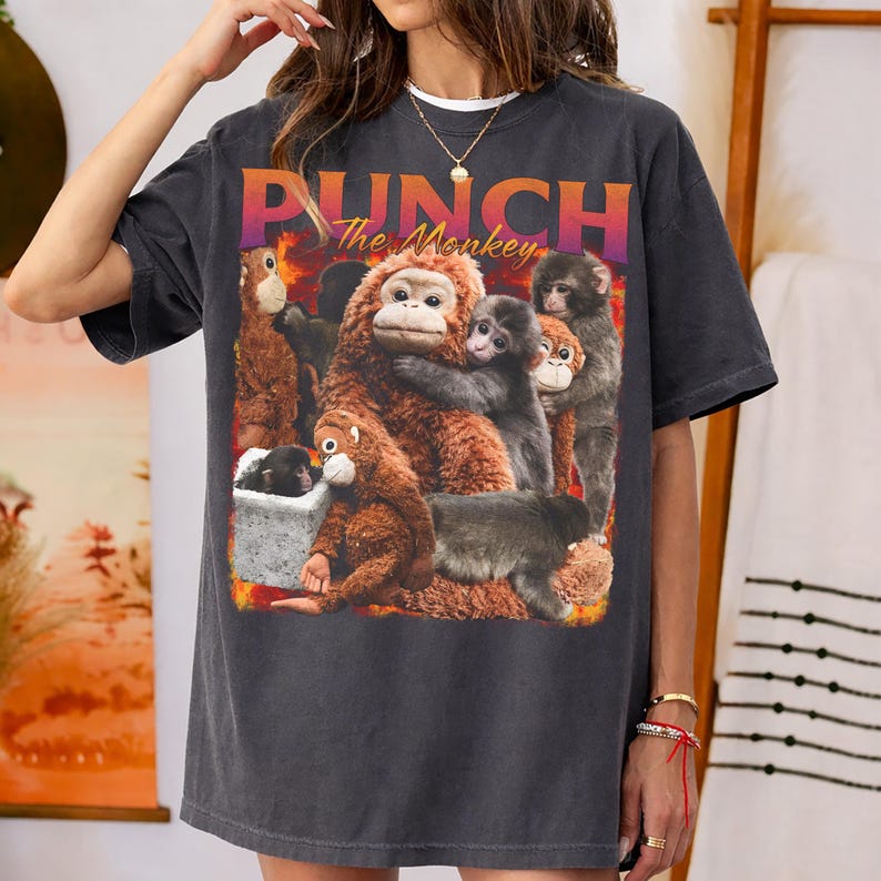 May include: Dark gray t-shirt featuring a graphic of several monkeys in various poses. The text "PUNCH The Monkey" is printed across the top in orange and purple. The shirt has a relaxed fit.