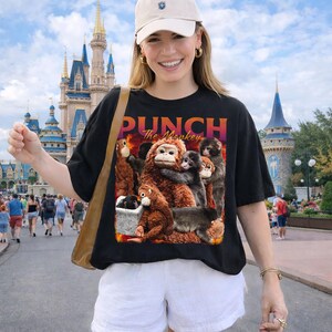 May include: Black t-shirt with a graphic of several monkeys and the text "PUNCH The Monkey" in orange and purple. The monkeys are various shades of brown and gray. The shirt is worn by a person.