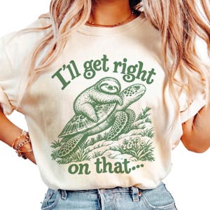 Könnte beinhalten: Ein cremefarbenes T-Shirt mit einer grünen Grafik, die ein Faultier auf dem Rücken einer Meeresschildkröte zeigt. Der Text "I'll get right on that..." ist in Grün über und unter der Illustration gedruckt, die auch kleine Grasbüschel enthält.