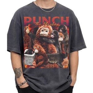May include: Dark gray t-shirt featuring a graphic of several monkeys with the text "PUNCH The Monkey" in a red and orange gradient. The monkeys are various shades of brown and black, creating a detailed illustration.