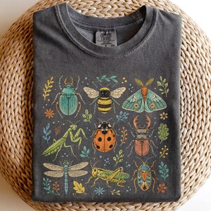 Op de afbeelding: Een antracietgrijs t-shirt met ronde hals en een grafische print van diverse insecten, waaronder een kever, bij, mot, bidsprinkhaan, lieveheersbeestje, vliegend hert, libel, sprinkhaan en wants, geaccentueerd met kleine botanische elementen.