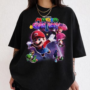Puede incluir: Camiseta negra con un gr&aacute;fico colorido del videojuego "The Super Mario Galaxy". El dise&ntilde;o incluye a Mario, Yoshi, la Princesa Peach y otros personajes sobre un fondo espacial. El texto "The Super Mario Galaxy" se muestra en la parte superior.