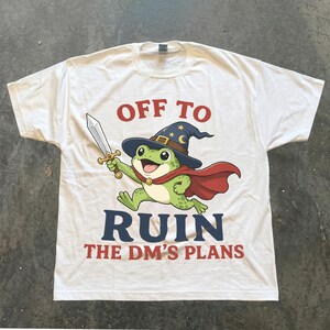 Może przedstawiać: Biały t-shirt z krótkim rękawem z grafiką zielonej żaby w niebieskim kapeluszu czarodzieja i czerwonej pelerynie, trzymającej miecz. Napis OFF TO RUIN THE DM'S PLANS jest nadrukowany czerwonymi i ciemnoniebieskimi literami na przodzie koszulki.