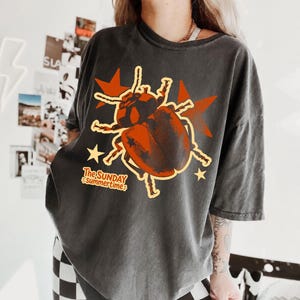 Può includere: Una t-shirt grigio antracite a maniche corte con una grande grafica di uno scarabeo marrone centrata sul petto. Il design include stelle arancioni e il testo The SUNDAY Summertime in un carattere arancione stilizzato.