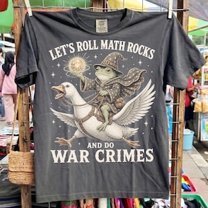 Può includere: Una t-shirt grigio antracite con la grafica di una rana vestita da mago, con tunica e cappello stellati, che cavalca un'oca bianca. Il testo recita LET'S ROLL MATH ROCKS AND DO WAR CRIMES, circondato da piccole stelle e un globo luminoso.