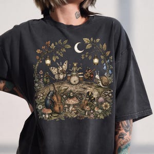Peut inclure: Un t-shirt gris anthracite orné d'une illustration détaillée représentant divers insectes jouant des instruments de musique dans une forêt sous un croissant de lune. Le dessin inclut un papillon de nuit, un scarabée, un escargot et une abeille.