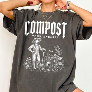 May include: A charcoal grey short-sleeved t-shirt featuring white screen-printed text that reads COMPOST YOUR ENEMIES. The graphic depicts a person in overalls and a hat holding a shovel, standing in a garden with plants and a skull in the soil.