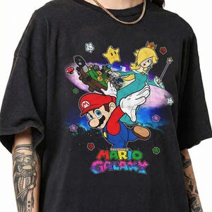 May include: Dark gray t-shirt featuring a colorful graphic of Mario and Princess Rosalina from Super Mario Galaxy. The design includes stars, a spaceship, and the game's logo in a vibrant, multi-colored font.