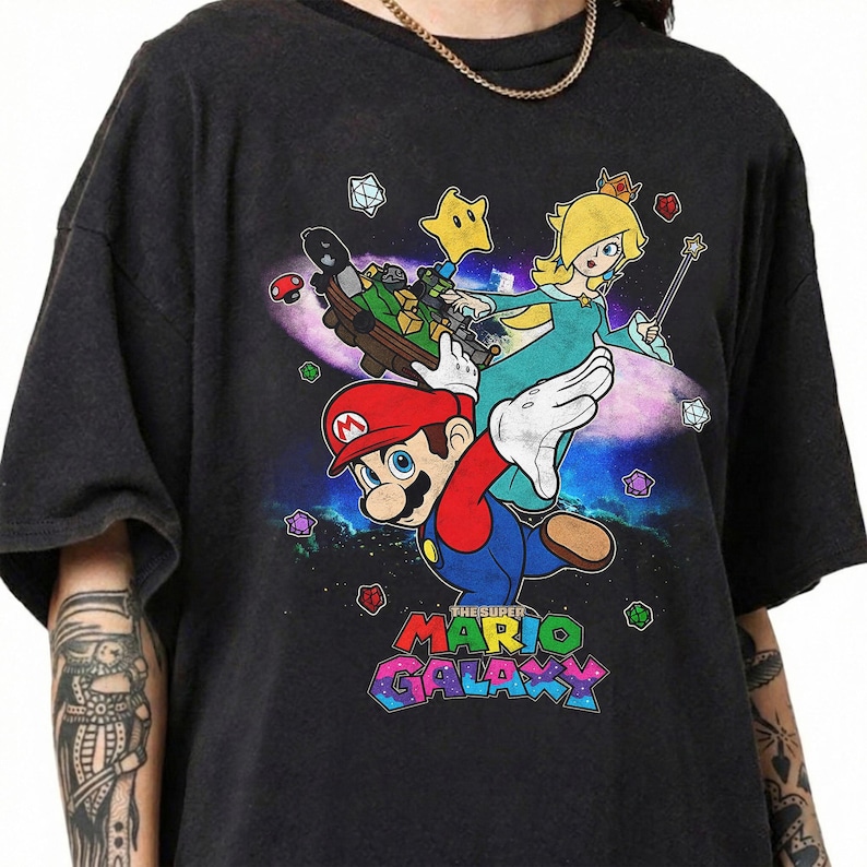 May include: Dark gray t-shirt featuring a colorful graphic of Mario and Princess Rosalina from Super Mario Galaxy. The design includes stars, a spaceship, and the game's logo in a vibrant, multi-colored font.