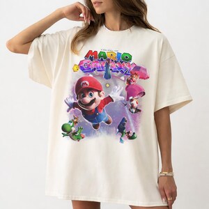 Puede incluir: Camiseta color crema con un gr&aacute;fico colorido de Mario, la Princesa Peach, Yoshi y otros personajes del videojuego "Mario Galaxy". El texto "MARIO GALAXY" se muestra en una fuente vibrante y multicolor sobre los personajes.