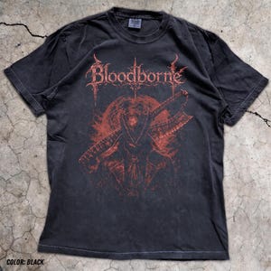 Puede incluir: Una camiseta negra de manga corta con un estampado gráfico rojo en el pecho. El diseño muestra el texto Bloodborne en una fuente estilizada sobre la ilustración de una figura con sombrero de ala ancha que sostiene una gran hoja dentada.