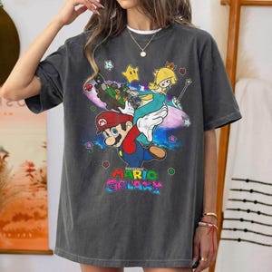 May include: Dark gray t-shirt featuring a colorful graphic of Mario, Rosalina, and other characters from the Super Mario Galaxy video game. The text "The Super Mario Galaxy" is displayed in a rainbow font at the bottom of the design.