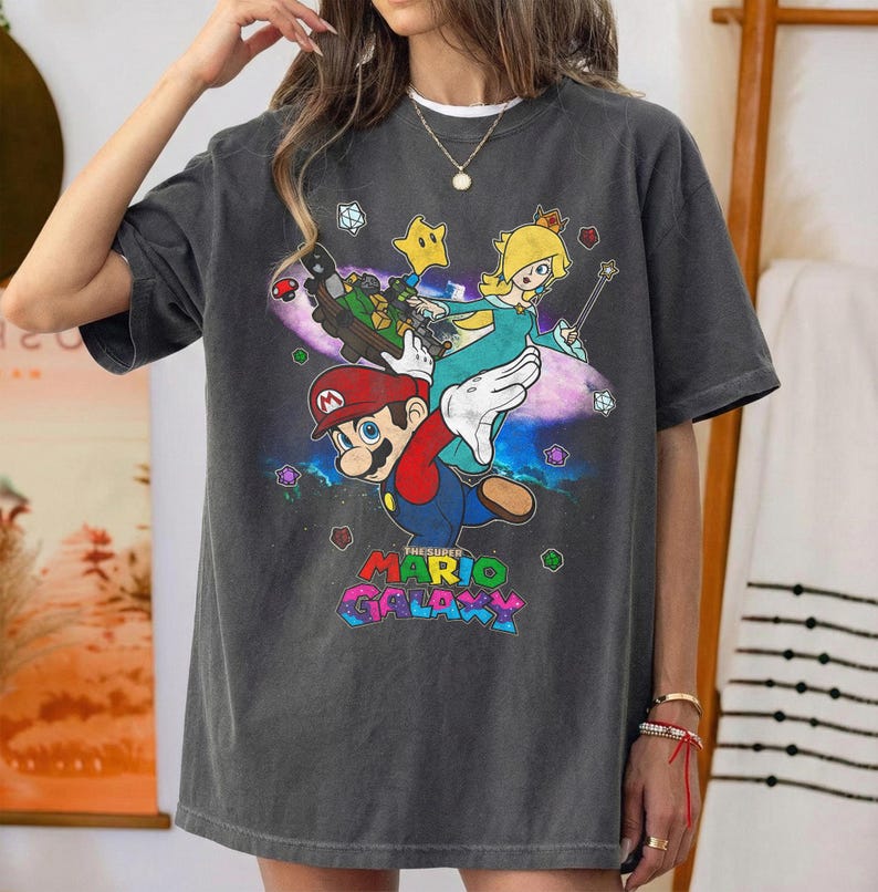 May include: Dark gray t-shirt featuring a colorful graphic of Mario, Rosalina, and other characters from the Super Mario Galaxy video game. The text "The Super Mario Galaxy" is displayed in a rainbow font at the bottom of the design.