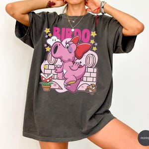 以下が含まれることがあります： ピンク色の漫画のキャラクターとピンク色の「BIRDO」の文字が特徴の濃いグレーのTシャツ。デザインには星、コイン、レンガの壁が含まれています。ゆったりとしたフィット感のTシャツです。