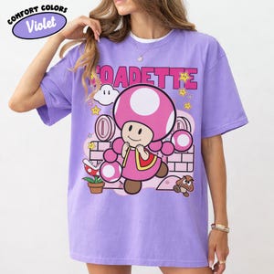 May include: Violet Comfort Colors t-shirt featuring a graphic of the character Toadette from the Super Mario video game series. The design includes the word "TOADETTE" in pink letters, along with stars, coins, and a brick wall background.