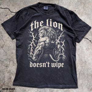 May include: A black short-sleeve t-shirt featuring a graphic of a muscular lion wearing a tank top, surrounded by lightning bolts. The text "the lion" appears above the lion, and the text "doesn't wipe" appears below the graphic in white.