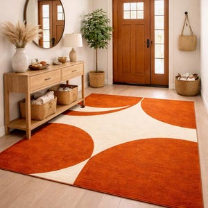 Könnte beinhalten: Ein Eingangsbereich mit einem orangefarbenen und cremefarbenen Teppich mit geometrischem Design. Der Raum umfasst eine Holzkonsolentisch, einen runden Spiegel und eine gewebte Pendelleuchte. Eine Topfpflanze und eine Holztür vervollständigen die Szene.