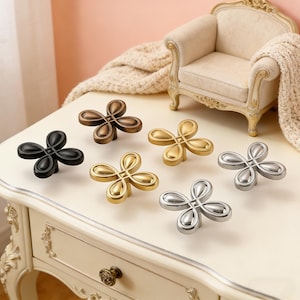 Puede incluir: Un conjunto de tiradores de armario con forma de flor en varios acabados metálicos, incluyendo negro, bronce, dorado y plateado. Los tiradores se exhiben sobre una superficie blanca, mostrando su diseño decorativo y su potencial para mejoras de mobiliario.