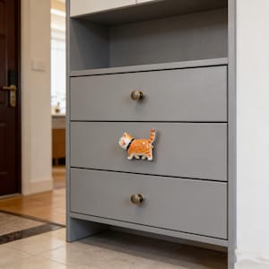 Peut inclure: Un meuble gris avec trois tiroirs et des boutons en laiton. Le tiroir du milieu présente un bouton en céramique en forme de chat tigré orange avec un collier noir. Le meuble fait partie d'un plus grand meuble de rangement.