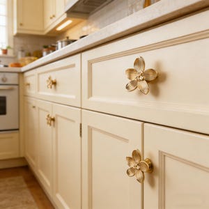 May include: Cream-colored kitchen cabinets with gold flower-shaped drawer pulls. The cabinet doors have a recessed panel design. The kitchen has a light-colored countertop and a white tile backsplash. The overall style is classic and elegant.