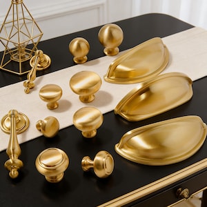May include: A collection of gold-toned cabinet hardware, including knobs and pulls, arranged on a black surface with a neutral runner. The hardware features various shapes, from spherical knobs to elongated cup pulls, all with a brushed finish. A decorative geometric candle holder is also present.