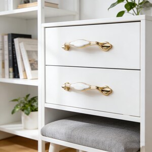 Peut inclure: Meuble blanc à deux tiroirs, chacun orné d'une poignée dorée avec un centre blanc de forme ovale. Les poignées présentent un design ornemental. Le meuble fait partie d'un ensemble avec un siège rembourré gris.