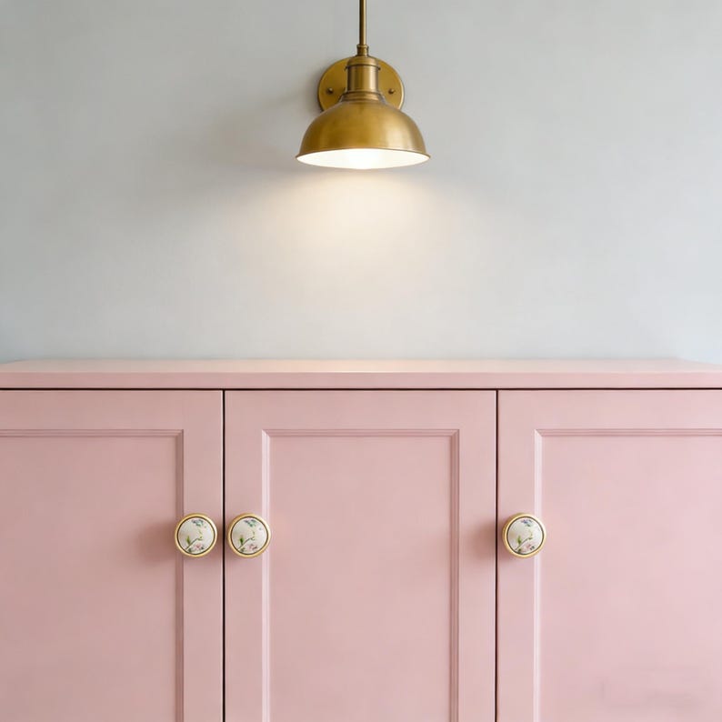 Peut inclure: Un meuble rose clair &agrave; trois portes avec des boutons &agrave; motifs floraux. Au-dessus, un luminaire mural en laiton avec un abat-jour en forme de d&ocirc;me. La lumi&egrave;re est allum&eacute;e, projetant une lueur chaleureuse.