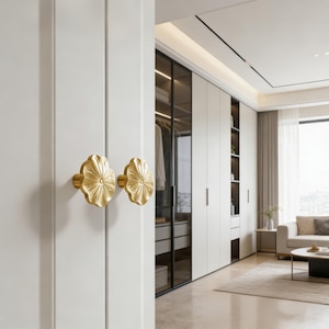 May include: Two gold-coloured, flower-shaped cabinet knobs are mounted on a white door. The knobs have a detailed petal design. The background shows a glimpse of a modern closet with white cabinets and glass doors, suggesting a home decor theme.