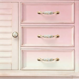Peut inclure: Un meuble rose clair avec trois tiroirs et une porte lat&eacute;rale. Les tiroirs ont des poign&eacute;es dor&eacute;es avec un motif floral. La porte de l'armoire a un bouton rond avec un motif floral assorti. L'armoire est peinte dans une palette de couleurs pastel douces.