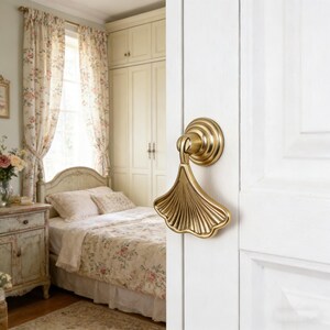 Może przedstawiać: Złoty uchwyt do drzwi w kształcie liścia miłorzębu japońskiego jest zamontowany na białych drzwiach. Uchwyt do drzwi ma okrągłą podstawę i szczegółowy, wachlarzowaty wzór liścia. Tło przedstawia sypialnię z kwiatowymi zasłonami i pościelą.
