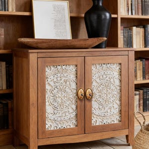May include: A wooden cabinet with two doors featuring intricate white carved designs. The cabinet has a natural wood finish and is topped with a wooden bowl, a framed picture, and a black vase. The cabinet doors have decorative oval handles.