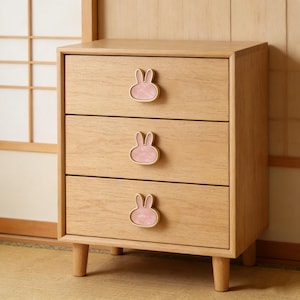 Puede incluir: Una mesita de noche de madera clara con tres cajones. Cada cajón presenta un tirador rosa en forma de conejito con un contorno dorado. La mesita tiene cuatro patas cortas y redondeadas.