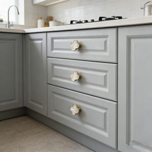 Puede incluir: Gabinetes de cocina gris claro con tiradores dorados y blancos en forma de trébol. Los gabinetes forman parte de una configuración de cocina en esquina, con una encimera blanca y un grifo de acero inoxidable.