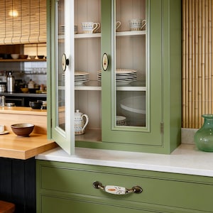 Op de afbeelding: Een groene keukenkast met glazen deuren, met witte borden, kopjes en een theepot. De kast heeft decoratieve handgrepen en staat tegen een bamboewand. De onderkasten hebben bijpassende groene laden met decoratieve handgrepen.