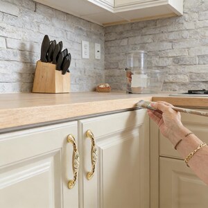 Puede incluir: Una cocina con encimera de madera clara y armarios color crema. Un bloque de cuchillos de madera contiene cuchillos negros. Una persona está pintando el borde de la encimera con un pincel. El salpicadero es un patrón de ladrillo gris.