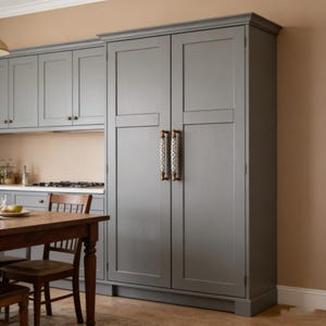 Puede incluir: Un gran armario de cocina gris con dos puertas y tiradores decorativos. El armario forma parte de un conjunto de cocina que incluye armarios superiores y una mesa de comedor de madera con sillas. La habitación tiene una paleta de colores neutros.