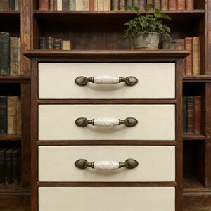 May include: A cream-colored wooden nightstand with four drawers. Each drawer has a brass-toned handle with a marble-like center. The nightstand is set against a backdrop of a bookshelf filled with books and a small potted plant.