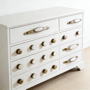 May include: A white dresser with eight drawers. The top drawers have brass and marble-look handles, while the others have brass knobs with white marble-look centers. The dresser is a classic piece of furniture.