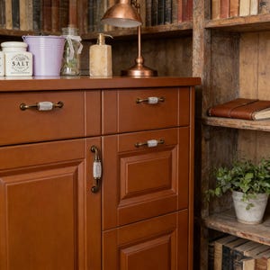 Peut inclure: Un meuble en bois brun avec des tiroirs et une porte, orné de quincaillerie décorative avec des accents blancs et gris. Une lampe de bureau cuivrée est posée sur le dessus, ainsi qu'un pot à sel, un seau violet et un bocal en verre. Une bibliothèque est en arrière-plan.