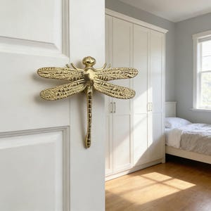 Op de afbeelding: Een goudkleurige deurklink in de vorm van een libel, gemonteerd op een witte deur. De klink is ingewikkeld gedetailleerd, met getextureerde vleugels en lichaam. Het libellenontwerp voegt een decoratief tintje toe aan de deur. De klink is waarschijnlijk van metaal.