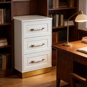 May include: A white three-drawer cabinet with gold accents. The cabinet has marble-look drawer pulls and a gold base. The cabinet is in a room with a desk, lamp, and bookshelves filled with books.