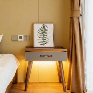 Puede incluir: Una mesita de noche moderna con un cajón gris y patas de madera. El cajón tiene un tirador decorativo. Una impresión enmarcada de helecho se encuentra encima. La mesita está iluminada desde abajo, creando un brillo cálido.